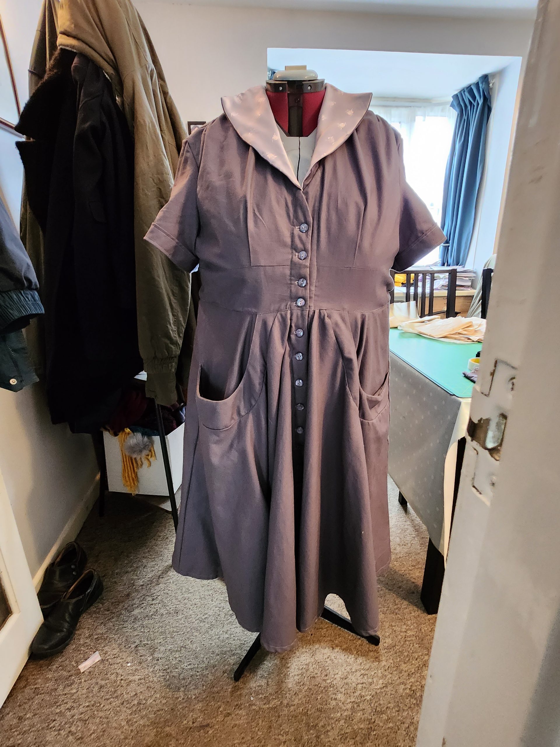 grey dress with pale purple lining on a dummy. It has a large collar with lining showing and pockets of unusually large size