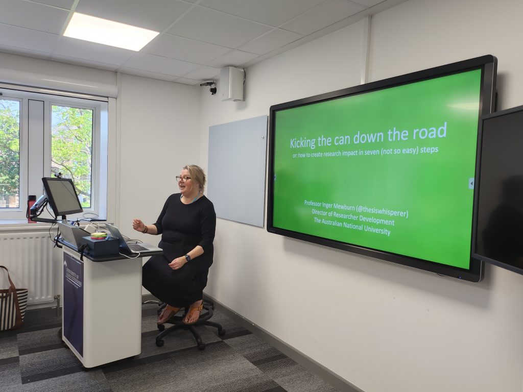 Photo of Inger Mewburn in front of a screen reading Kicking the can down the road or how to create research impact in seven (not so easy) steps.
